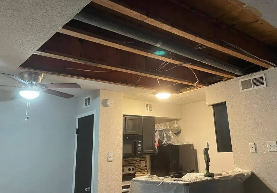 Apartment living room with a large section of ceiling removed, joists, duct and wiring exposed.