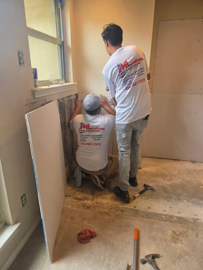 Two ML crew members in branded white shirts fitting new drywall under a bedroom window.