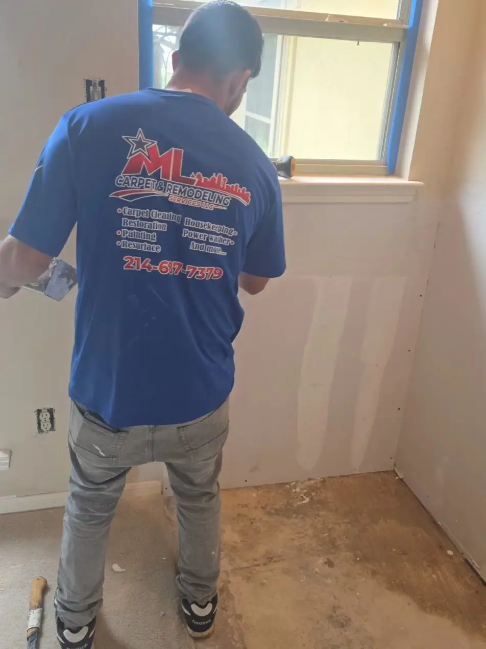 Crew member in a blue ML shirt mudding new drywall seams under a window.