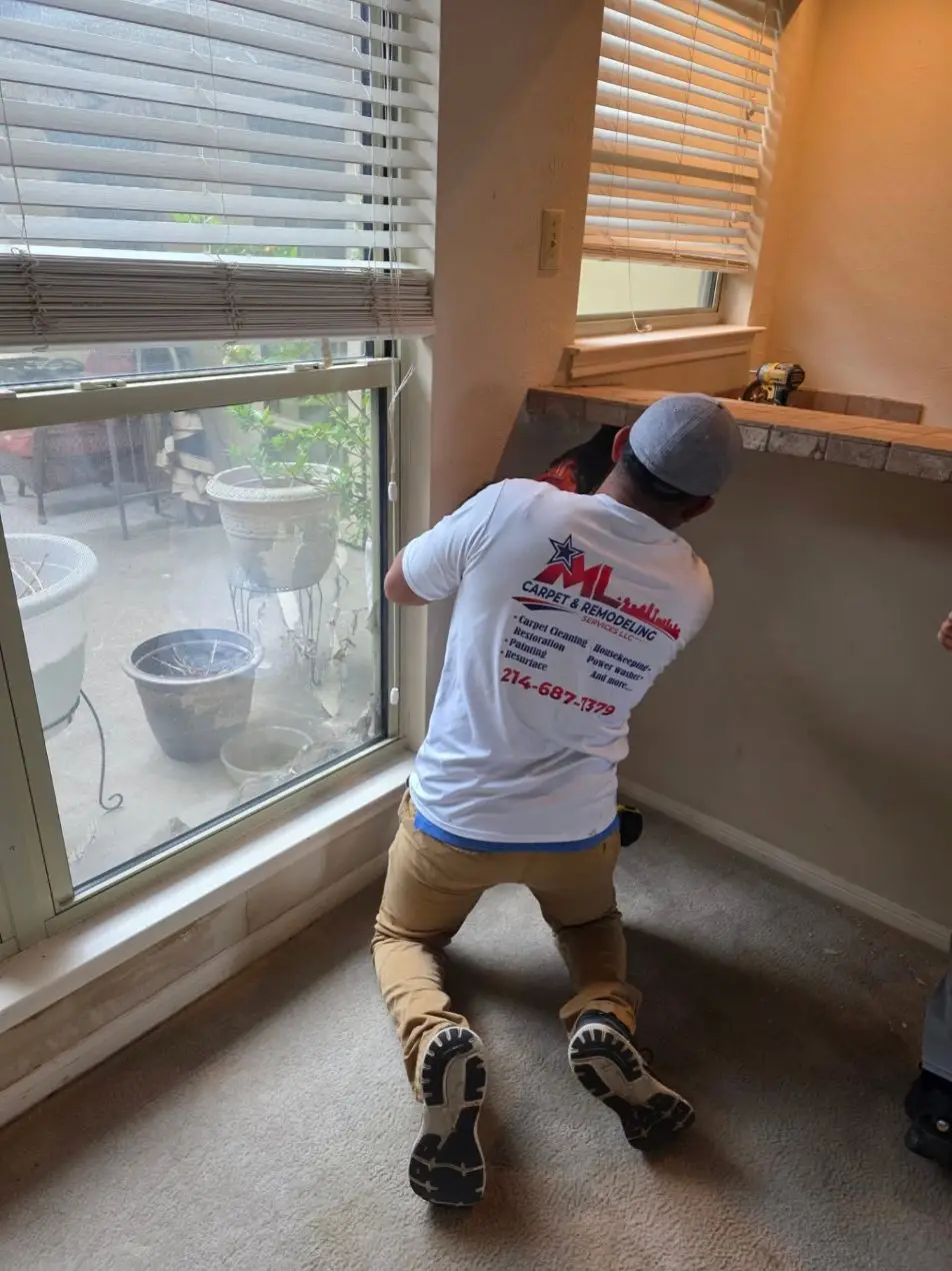 Crew member in a white ML shirt kneeling to cut out water-damaged drywall under a window.